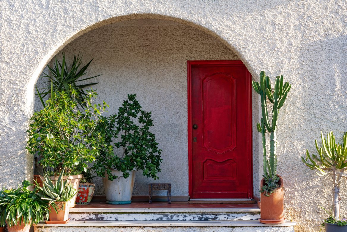 A red front door under a stucco arch, framed by potted palms and succulents