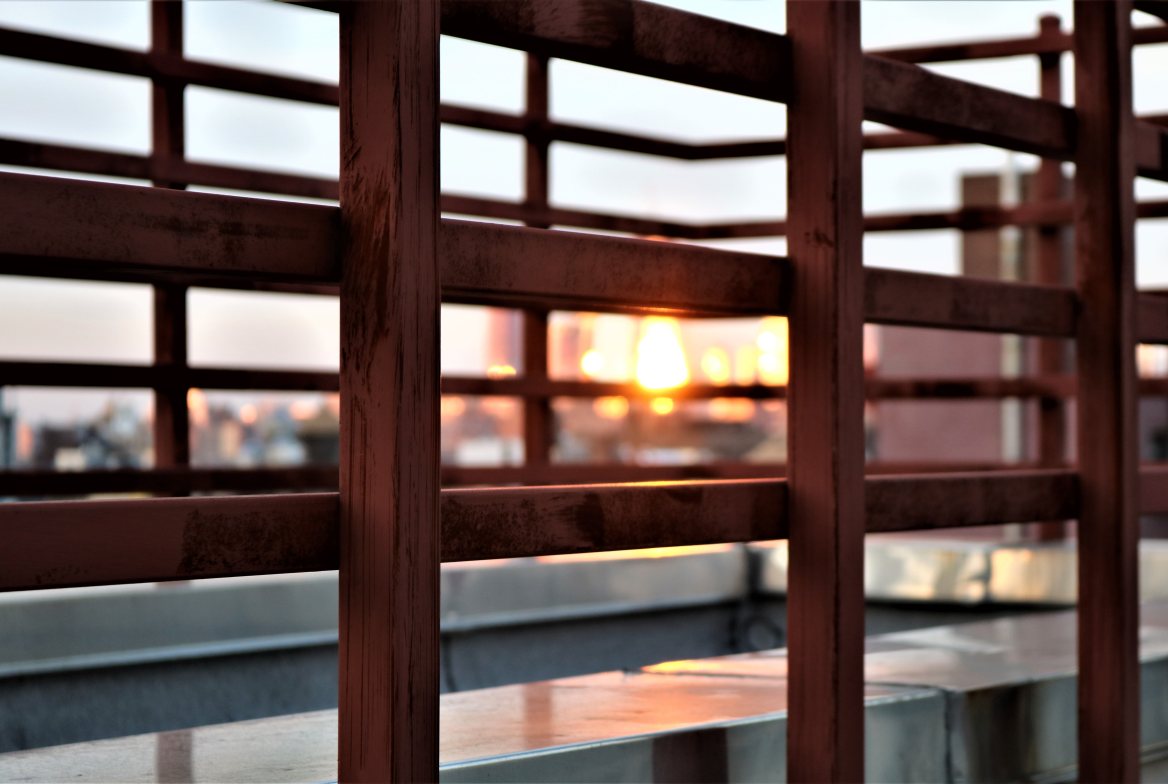 The sun setting behind a balcony&rsquo;s wooden rails, city softly out of focus