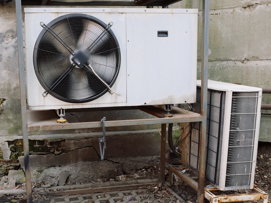 An old, rusted outdoor AC unit on a metal stand beside a smaller condenser