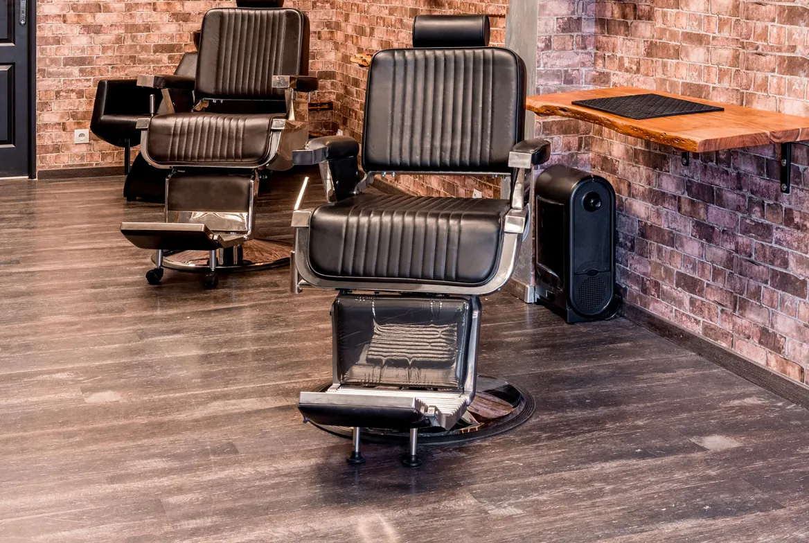 Two empty leather barber chairs on a wood floor beside a brick wall