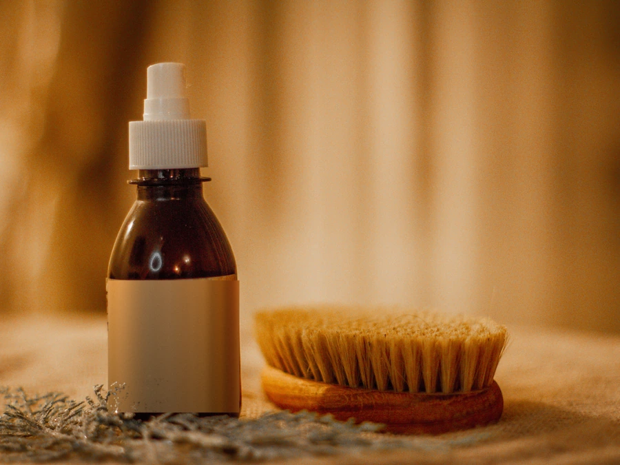 A bottle of beard oil beside a wooden beard brush in warm light