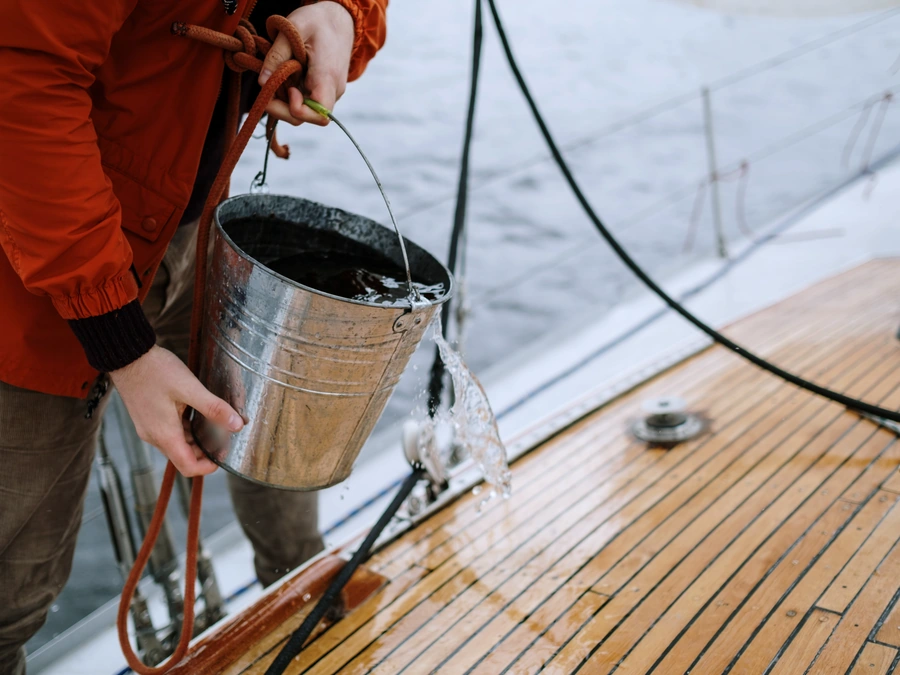 A detailer's hands pouring water from a steel bucket onto a teak boat deck