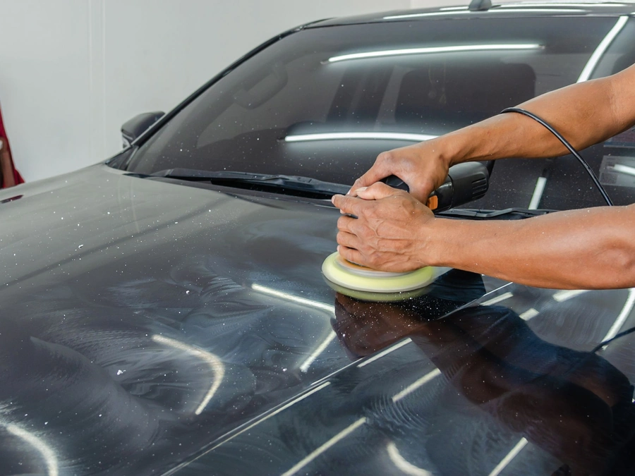 A detailer's hands running a dual-action polisher over a black car hood marked with swirl scratches