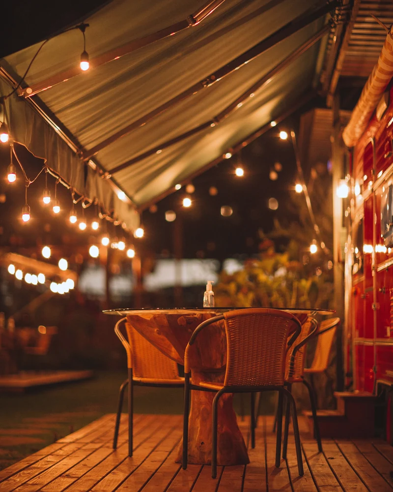 A covered patio at night with string lights, a striped awning and wicker chairs around a table