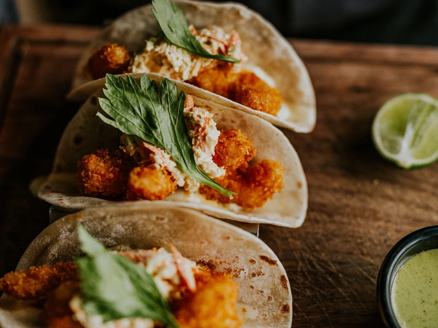 Three crispy fish tacos topped with slaw and herbs, with a lime half on the side